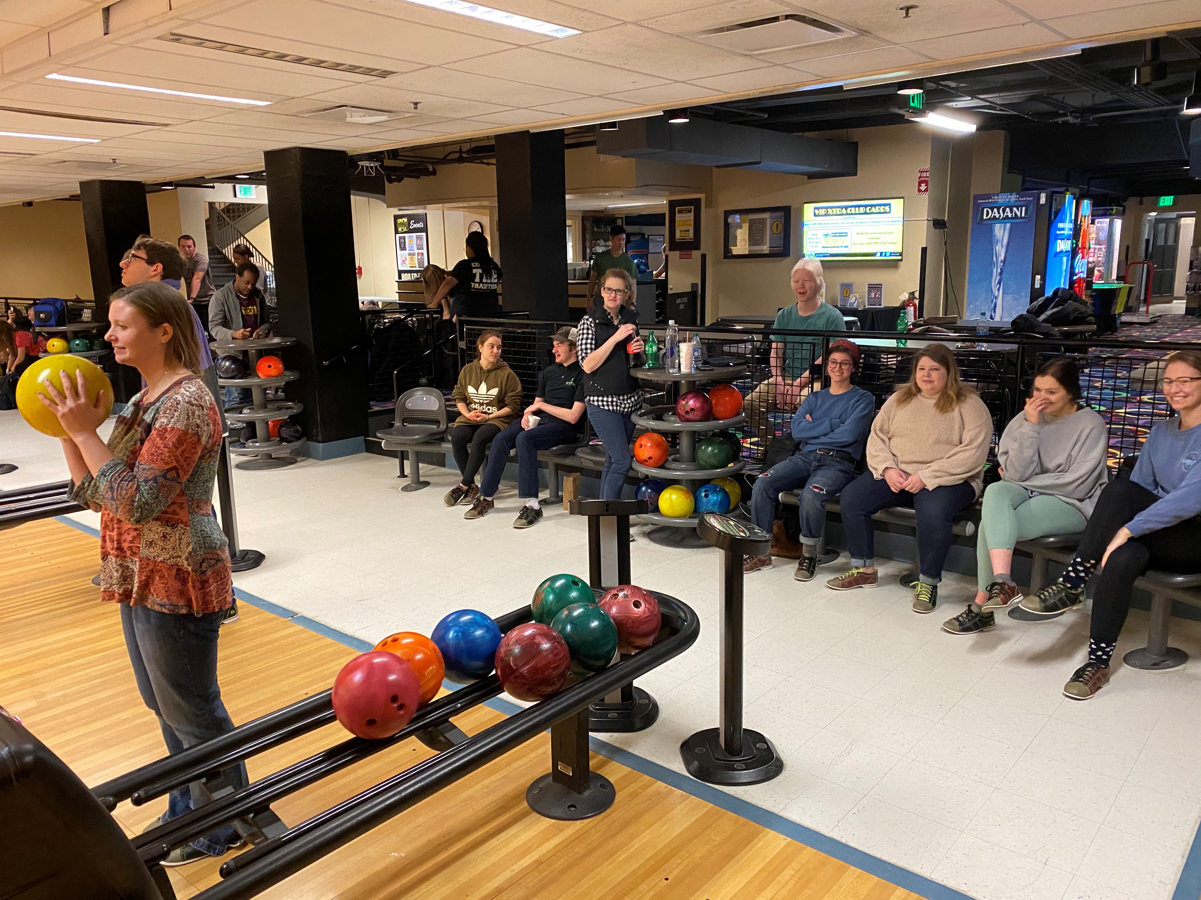 Picture of a woman with a bowling ball, preparing to take her shot, while others look on from the background at the DRC Peer Mentor Program Bowling Night.